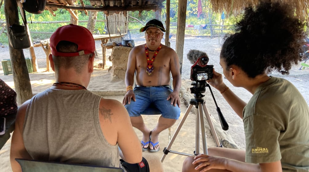 Two journalists in the foreground record a video interview with an Indigenous leader (in the background) using a tripod-mounted camera