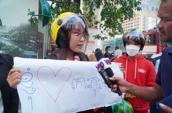 A young woman in a colorful helmet holds up a poster with a heart and speaks into a microphone