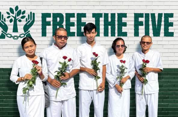 Five people dressed in white, holding red roses, standing in front of a wall that reads “Free the Five”