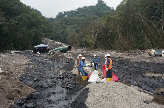 A group of oil workers work between two streams of black oil that have spilled down a boulder-covered mountainside in the rainforest. In the background, a section of pipe from the oil pipeline can be seen.