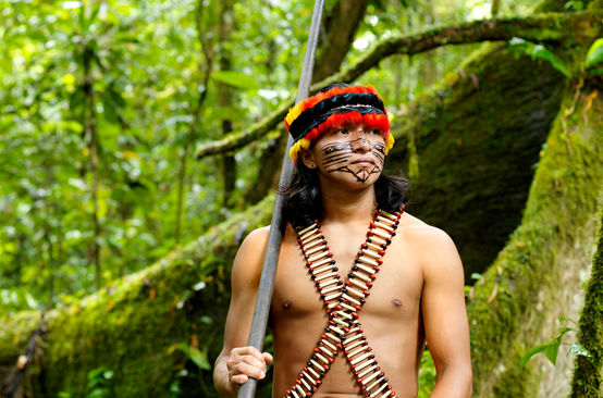 An indigenous man wearing feathers and a painted face standing in the forest with a blowpipe