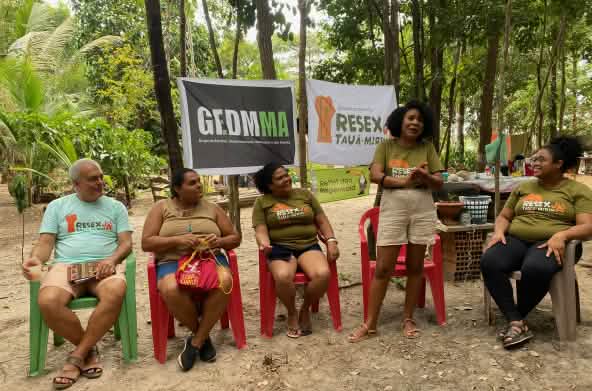 A woman speaks surrounded by four people sitting on chairs under trees and in front of three banners stretched across the tree trunks