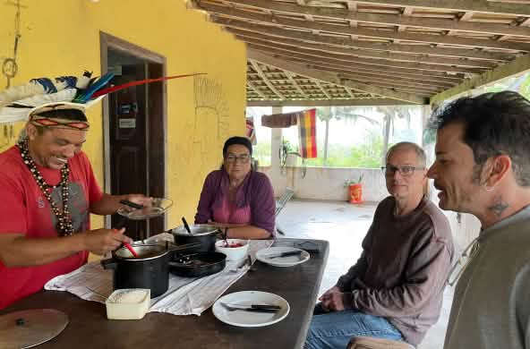 Mady Pataxó, adorned with a feather headdress, his mother, and Rainforest Rescue activists Felipe Sabrina and Klaus Schenck sit around a wooden table set for lunch.
