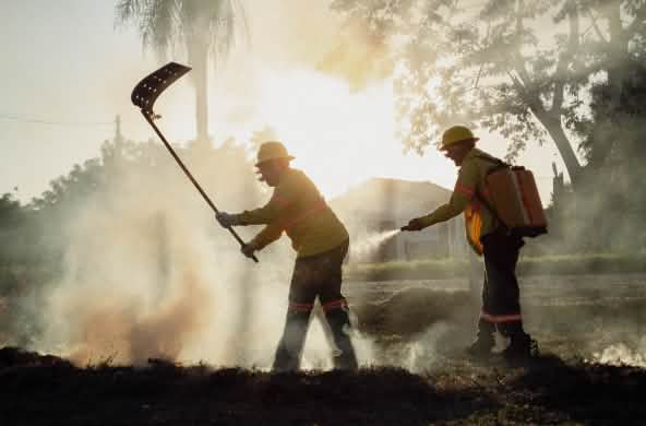 Two men with a fire beater and a water backpack with a hand-held spray nozzle surrounded by smoke