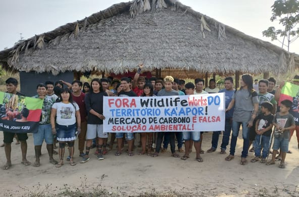 A group of Indigenous people with protest banners in front of a hut thatched with palm fronds