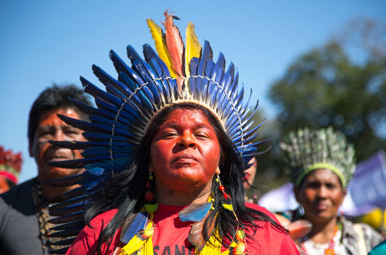 An indigenous woman adorned with a feathered crown in front of two other indigenous people