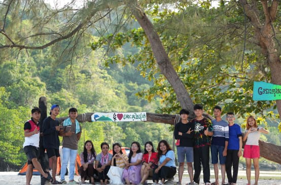 A group of young people sitting and standing in front of a tree on the beach