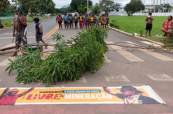 Indigenous people are blocking a road, with a line of people in the background and branches and a protest banner in the foreground