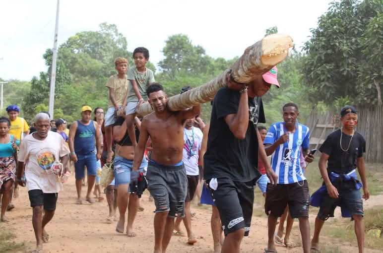 Men and women carry a log on their shoulders. Two children sit on top of it.