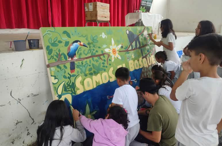 About ten children are painting a large picture of plants and animals, which is leaning against a wall