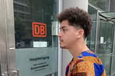 A young man stands with an envelope in front of a glass door with the inscription DB Main Entrance, Corporate Headquarters