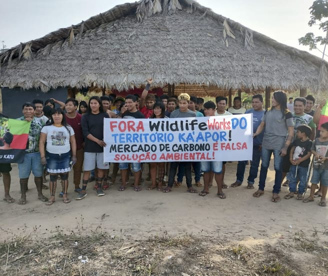 A group of Indigenous people with protest banners in front of a hut thatched with palm fronds