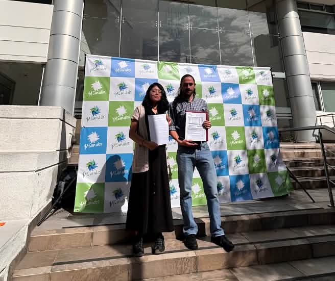 A woman and a man standing in front of a building entrance next to a banner reading “YASunidos,” holding documents in their hands