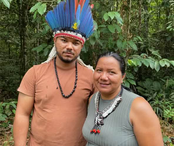 Portrait of an Indigenous woman wearing a feather necklace on the right and a young Indigenous man wearing a feather headdress on the left