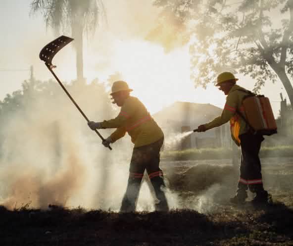 Two men with a fire beater and a water backpack with a hand-held spray nozzle surrounded by smoke