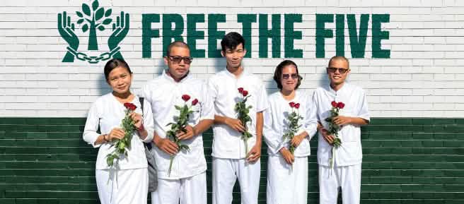 Five people dressed in white, holding red roses, standing in front of a wall that reads “Free the Five”