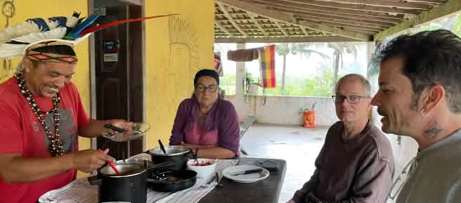 Mady Pataxó, adorned with a feather headdress, his mother, and Rainforest Rescue activists Felipe Sabrina and Klaus Schenck sit around a wooden table set for lunch.