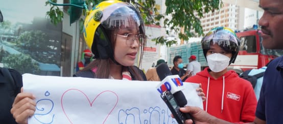 A young woman in a colorful helmet holds up a poster with a heart and speaks into a microphone