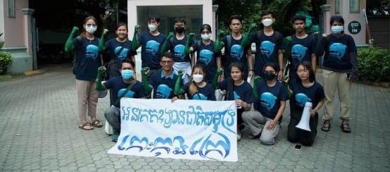 A group of 15 young people standing and kneeling in front of the Ministry of Tourism building, holding a banner to the camera. It reads “Koh Kong Island must become a marine national park”