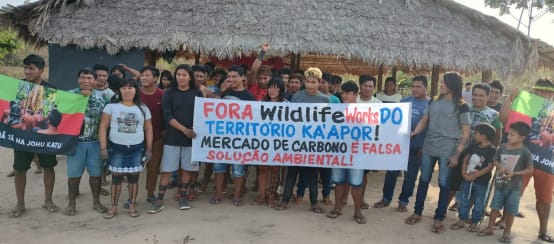 A group of Indigenous people with protest banners in front of a hut thatched with palm fronds
