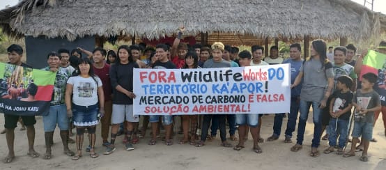 Ka’apor protest against carbon traders in their territory. Inscription on the banner: Wildlife Works out of Ka’apor territory! The carbon market is a false environmental solution! (© TUXA TA PAME) A group of Indigenous people with protest banners in front of a hut thatched with palm fronds