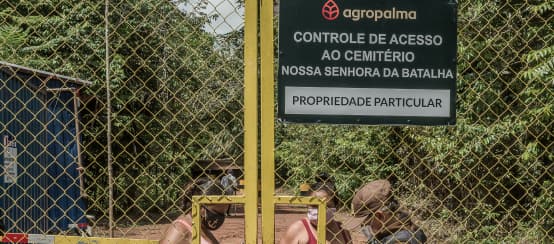 Local people cannot visit their own cemetery without dealing with Agropalma’s fences and guard posts (© Avispa Mídia) Two women behind a high barred gate are being checked by a guard. Inscription on the sign: Agropalma. Access control for Nossa Senhora da Batalha cemetery. Private property