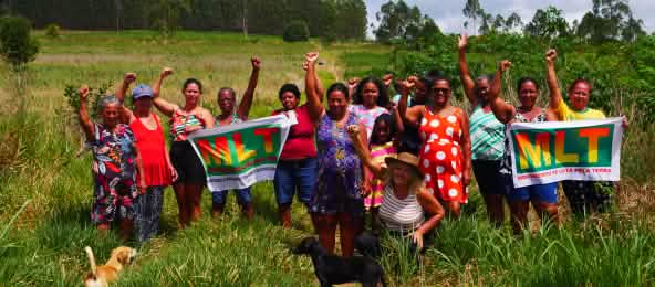 A group of 15 women is protesting with their fists raised and two banners bearing the letters MLT