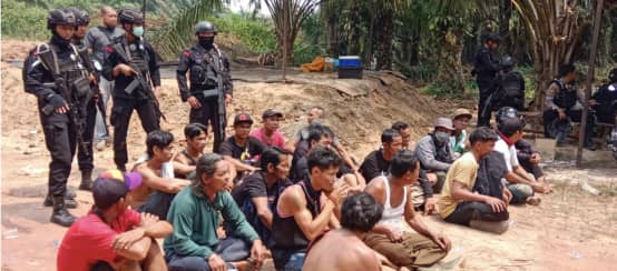 Detained Dayak people sit on the ground, armed police officers behind them