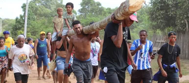 Men and women carry a log on their shoulders. Two children sit on top of it.