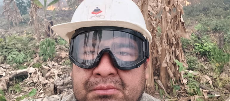 A man looks into the camera wearing a helmet and goggles while bushes burn and smoke rises in the background
