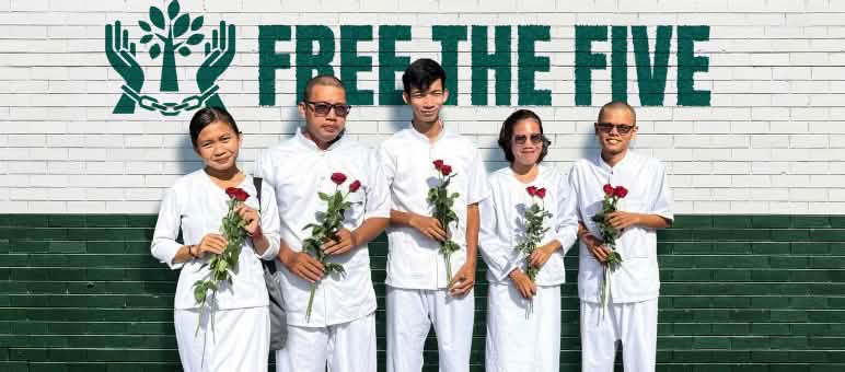 Five people dressed in white, holding red roses, standing in front of a wall that reads “Free the Five”