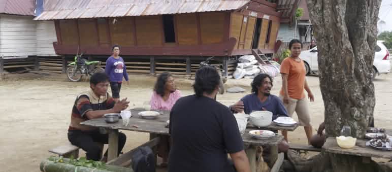 Four people sitting at a table outside, with a Batak house in the background