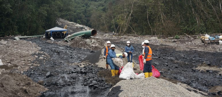 A group of oil workers work between two streams of black oil that have spilled down a boulder-covered mountainside in the rainforest. In the background, a section of pipe from the oil pipeline can be seen.