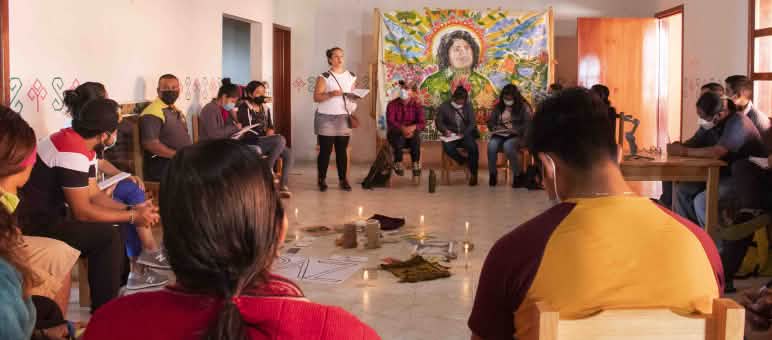 People are sitting in a circle, participating in a workshop. In the center are lit candles and a piece of paper with the letters PAZ written on it. A portrait of Berta Cáceres hangs on the wall