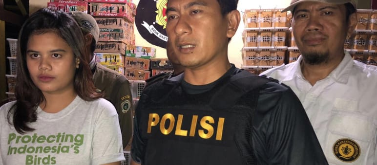 Young woman wearing Flight t-shirt and police officer in a warehouse with crates of birds