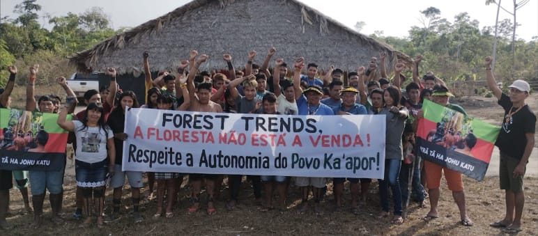 A group of Indigenous people with protest banners in front of a hut covered with palm fronds