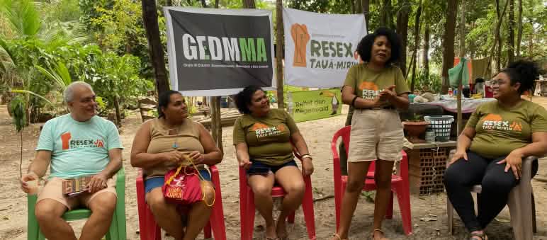 A woman speaks surrounded by four people sitting on chairs under trees and in front of three banners stretched across the tree trunks