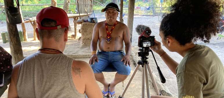 Two journalists in the foreground record a video interview with an Indigenous leader (in the background) using a tripod-mounted camera