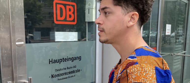 Mikaell from the Brazilian organization Justiça nos Trilhos in front of the DB headquarters in Berlin A young man stands with an envelope in front of a glass door with the inscription DB Main Entrance, Corporate Headquarters