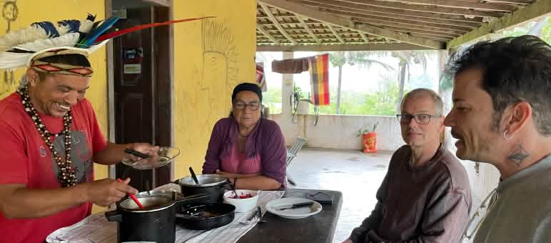 Mady Pataxó, adorned with a feather headdress, his mother, and Rainforest Rescue activists Felipe Sabrina and Klaus Schenck sit around a wooden table set for lunch.
