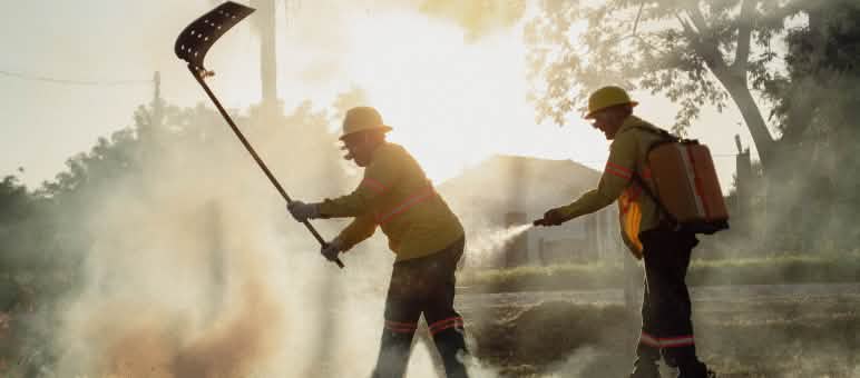 Two men with a fire beater and a water backpack with a hand-held spray nozzle surrounded by smoke