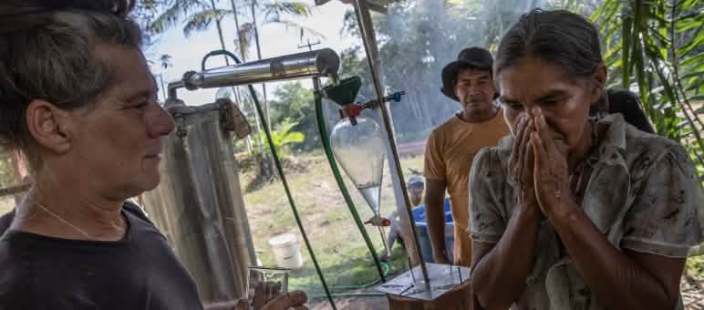 An Indigenous woman standing beside a steam distillation unit smells essential oils that the instructor has dripped onto her hand