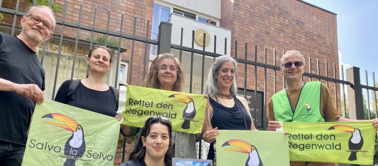 Six people standing in front of the Cambodian embassy compound's fence, holding banners with the organization’s toucan logo and the texts Rettet den Regenwald and Salva la Selva