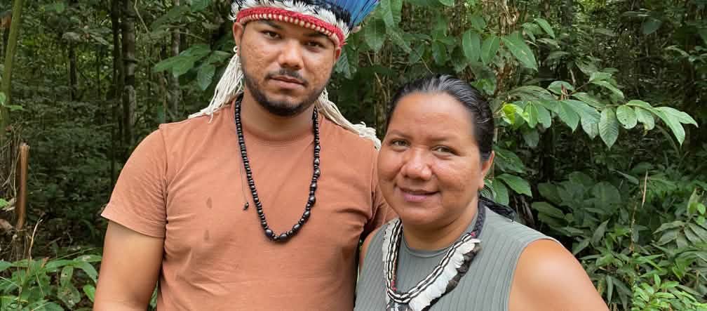 Portrait of an Indigenous woman wearing a feather necklace on the right and a young Indigenous man wearing a feather headdress on the left