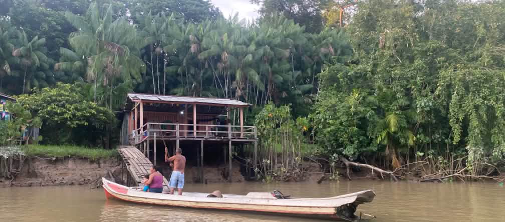 Two people in a wooden boat in front of a wooden shack in on a riverbank
