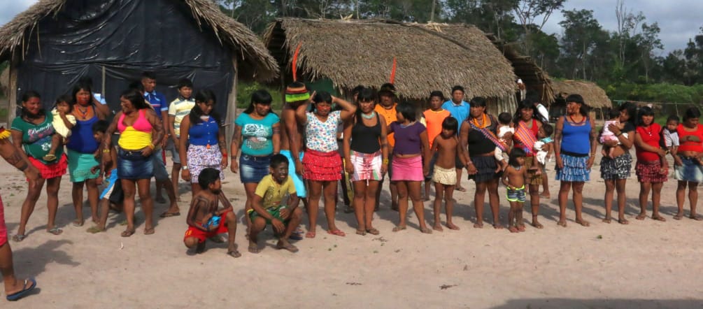 Indigenous Ka'apor standing in a double row in front of two huts in the forest