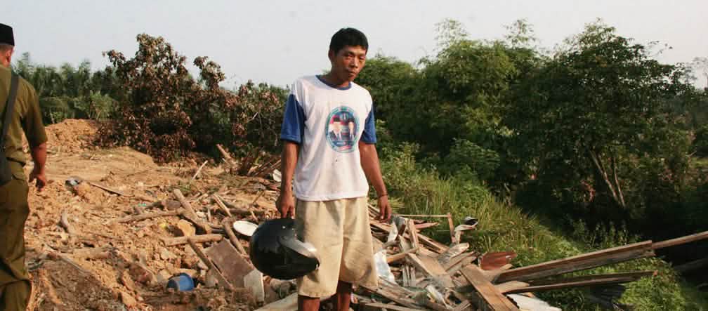 A farmer stands in front of the ruins of his village