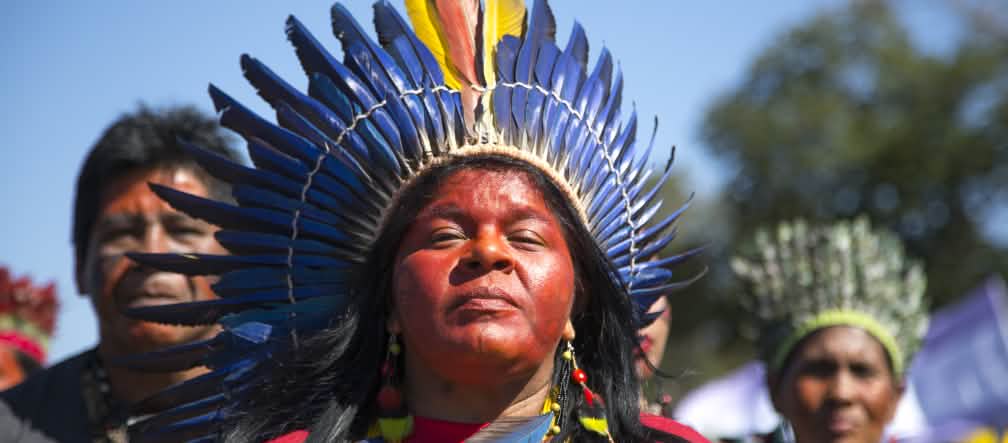 An indigenous woman adorned with a feathered crown in front of two other indigenous people