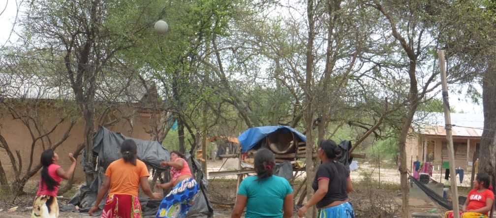 Five indigenous women play a ball game between trees and wooden houses