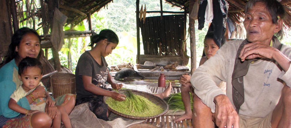 A man, three women and a small boy sit in an open hut, a woman sorts rice in a shallow bowl